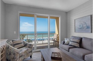 A room with a balcony overlooking a beach, a sofa, a chair, and a coffee table.