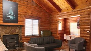 L’intérieur d’une cabane en rondins, avec une cheminée en pierre, un canapé, deux fauteuils et un tapis à motifs.