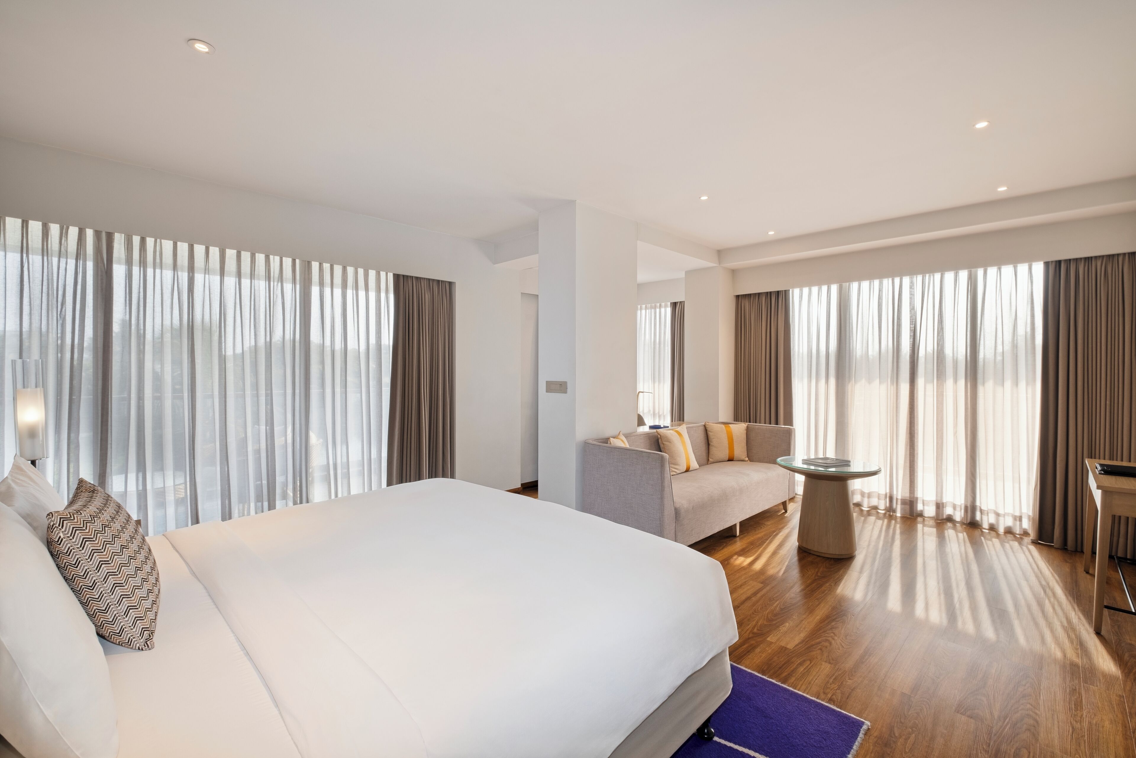 Una habitación de hotel moderna con una cama grande, un sofá, una mesita y vistas a través de cortinas translúcidas.