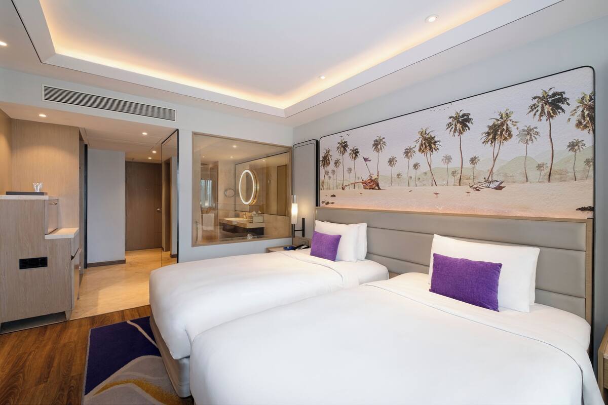 A hotel room with two beds, a large painting of a beach scene, a minibar, and a bathroom with a mirror and sink.