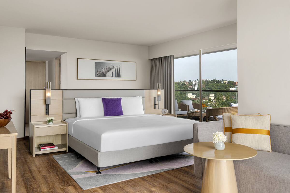 A modern hotel room with a large bed, a sofa, a small table, and a view of the city.