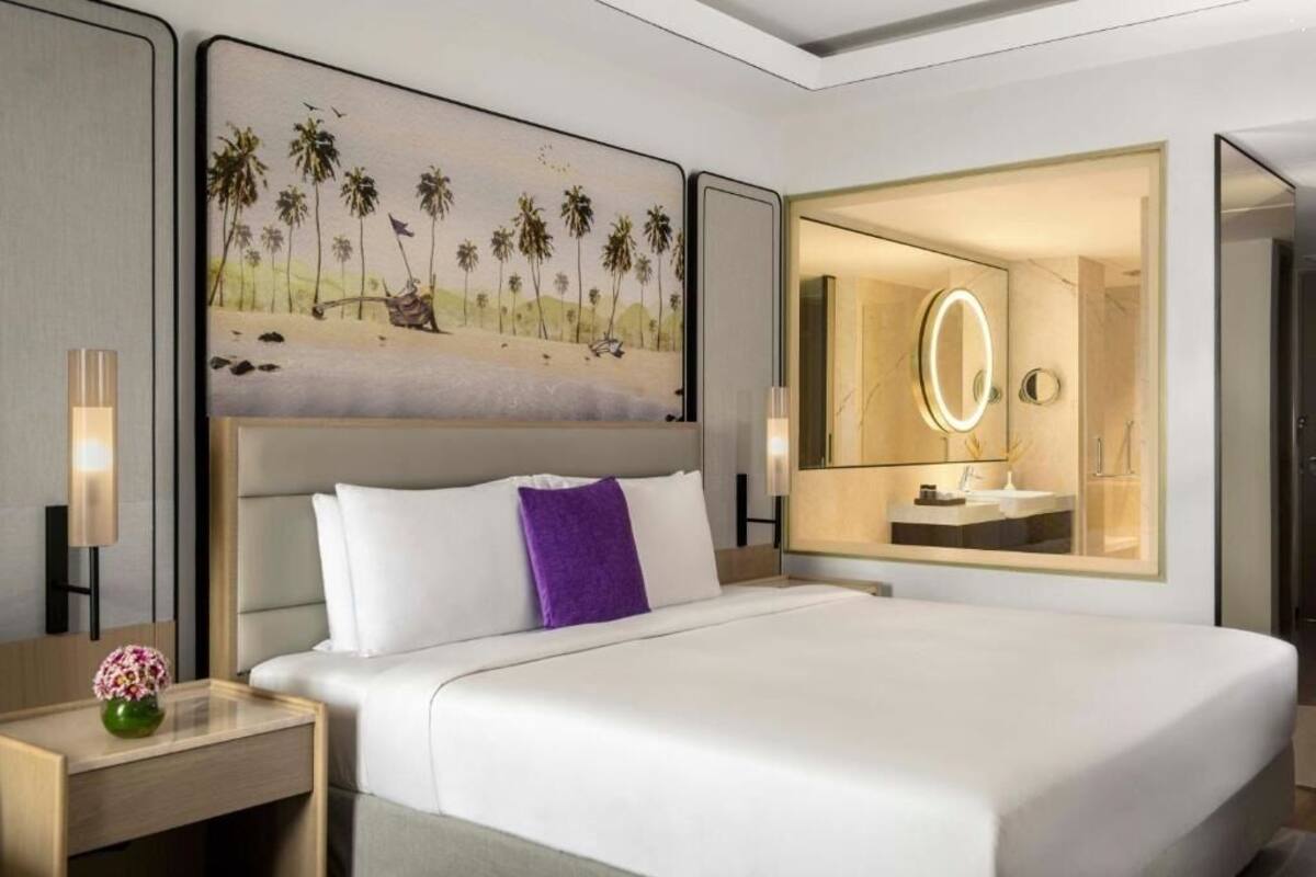 A hotel room with a bed, a nightstand with a vase, a painting of a beach scene, and a bathroom with a mirror.