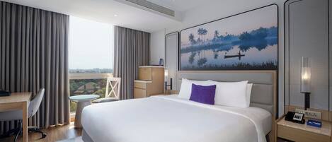 A modern hotel room with a large bed, a desk, a chair, a small table, and a painting on the wall.