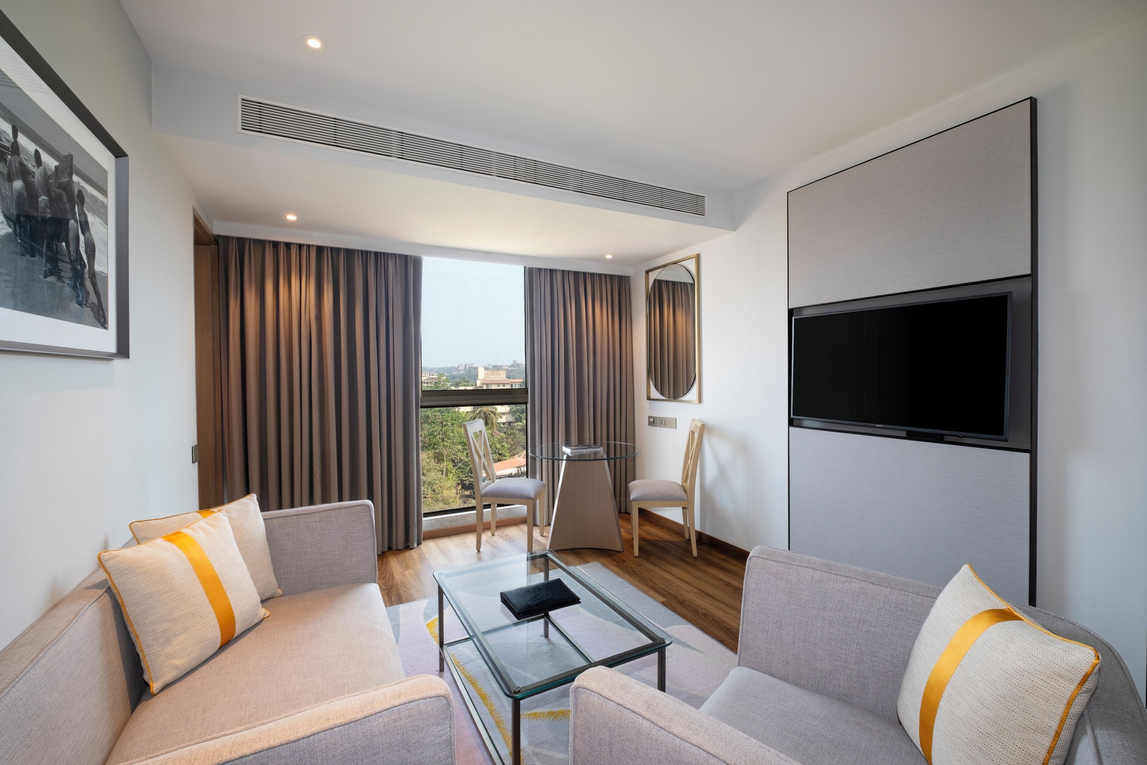 A modern hotel room with a sofa set, a glass coffee table, a flat-screen TV mounted on the wall, and a large window with curtains.