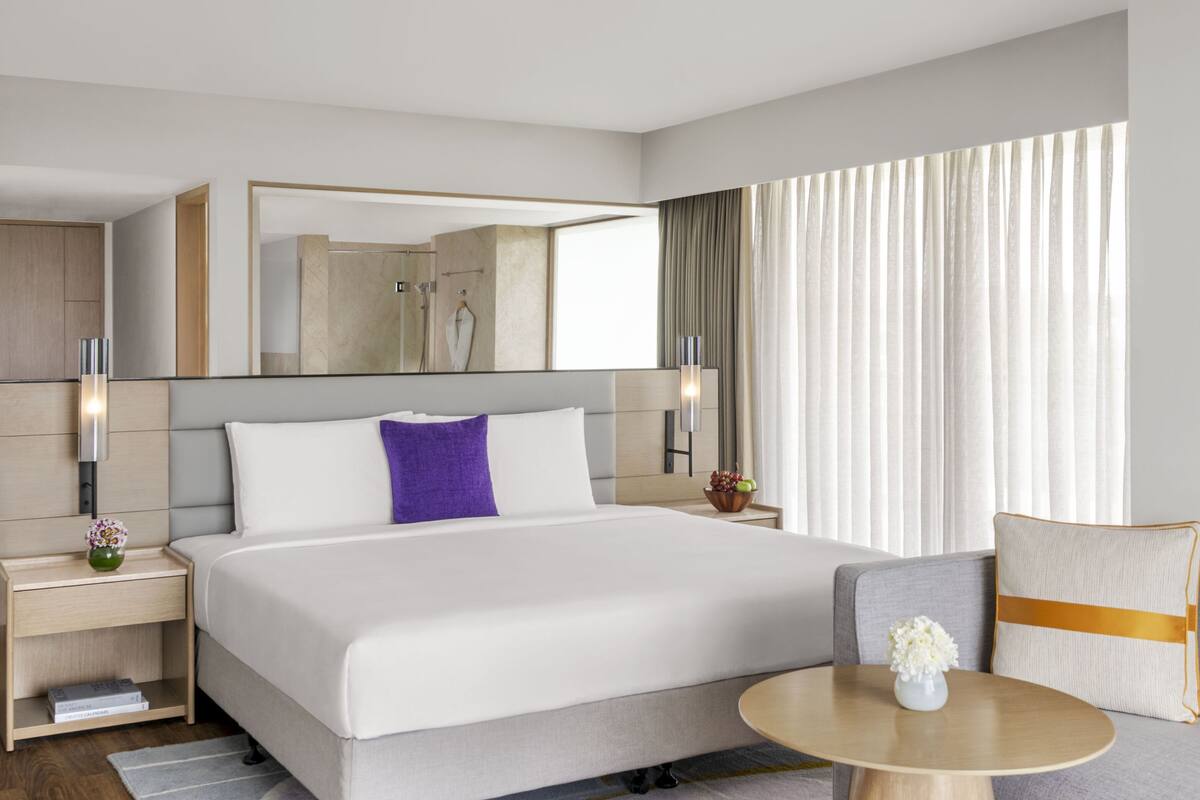 A modern hotel room with a large bed, a bedside table with a lamp, a small table with a vase, and a sofa.