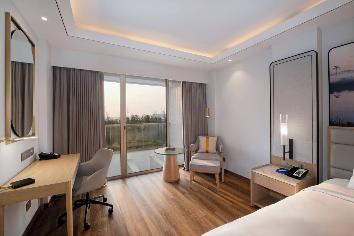 A modern hotel room with a wooden desk, a comfortable armchair, a small bedside table with a lamp, and a large window offering a view of the outdoors.