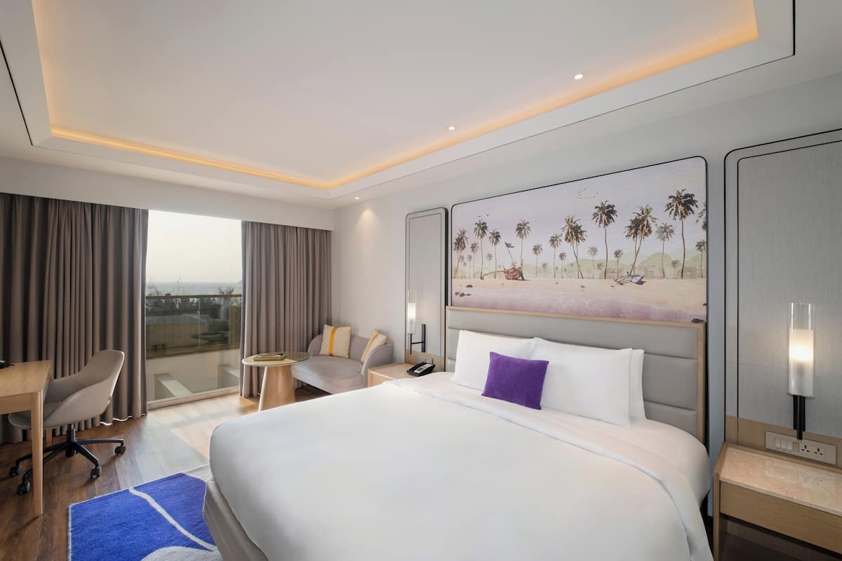 A hotel room with a large bed, a desk, a chair, a small table, and a painting of palm trees.