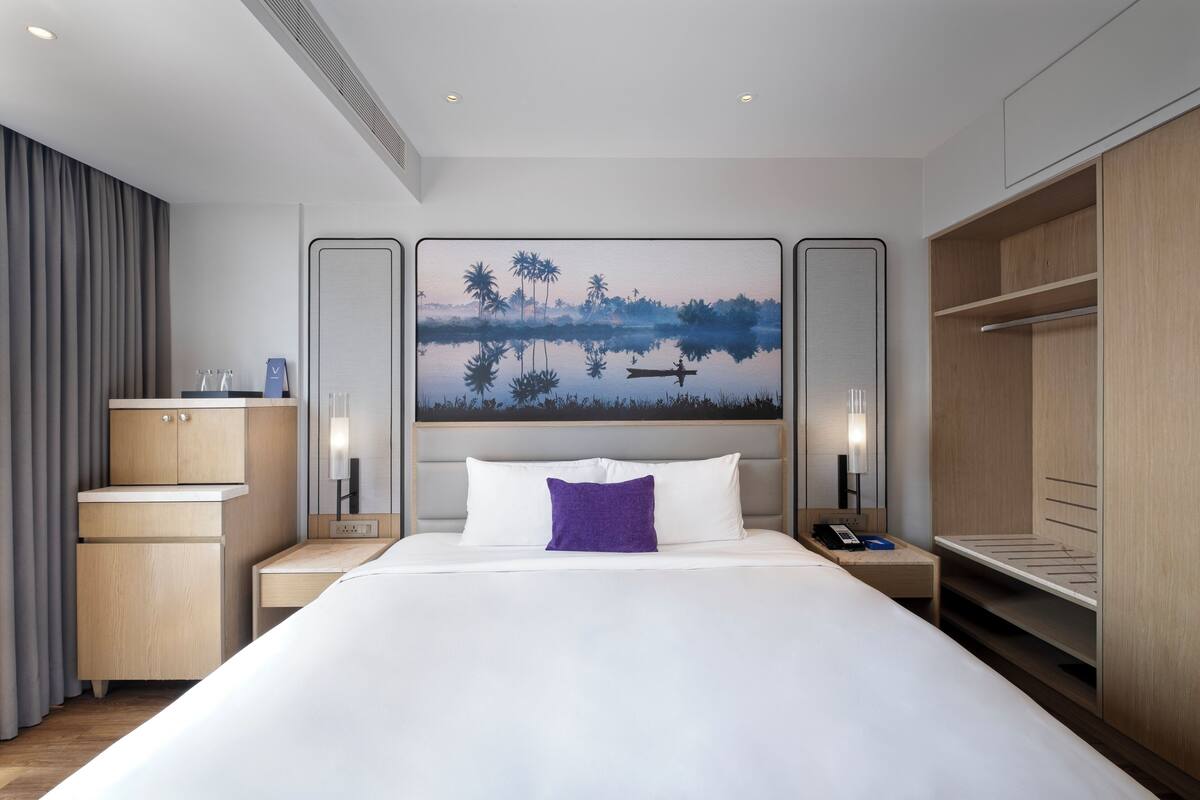 A modern hotel room with a large bed, a nightstand with a phone, a desk, and a wardrobe.