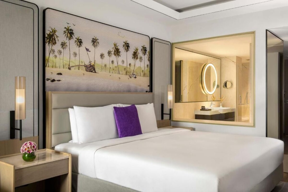 A hotel room with a bed, a nightstand with a vase, a painting of palm trees, and a bathroom with a mirror.