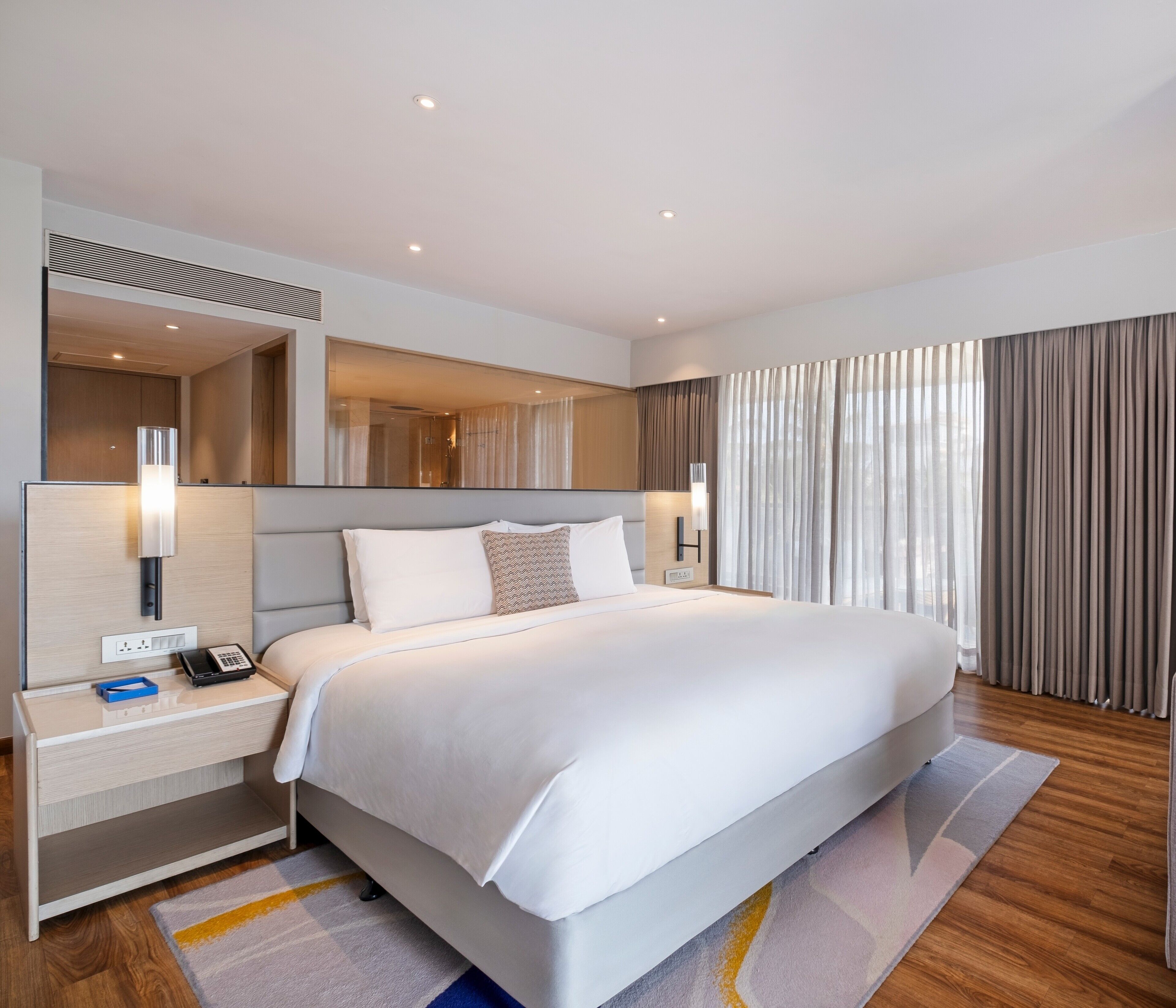 A modern hotel room with a large bed, a bedside table with a phone, a mirror, and a view of the outside through sheer curtains.
