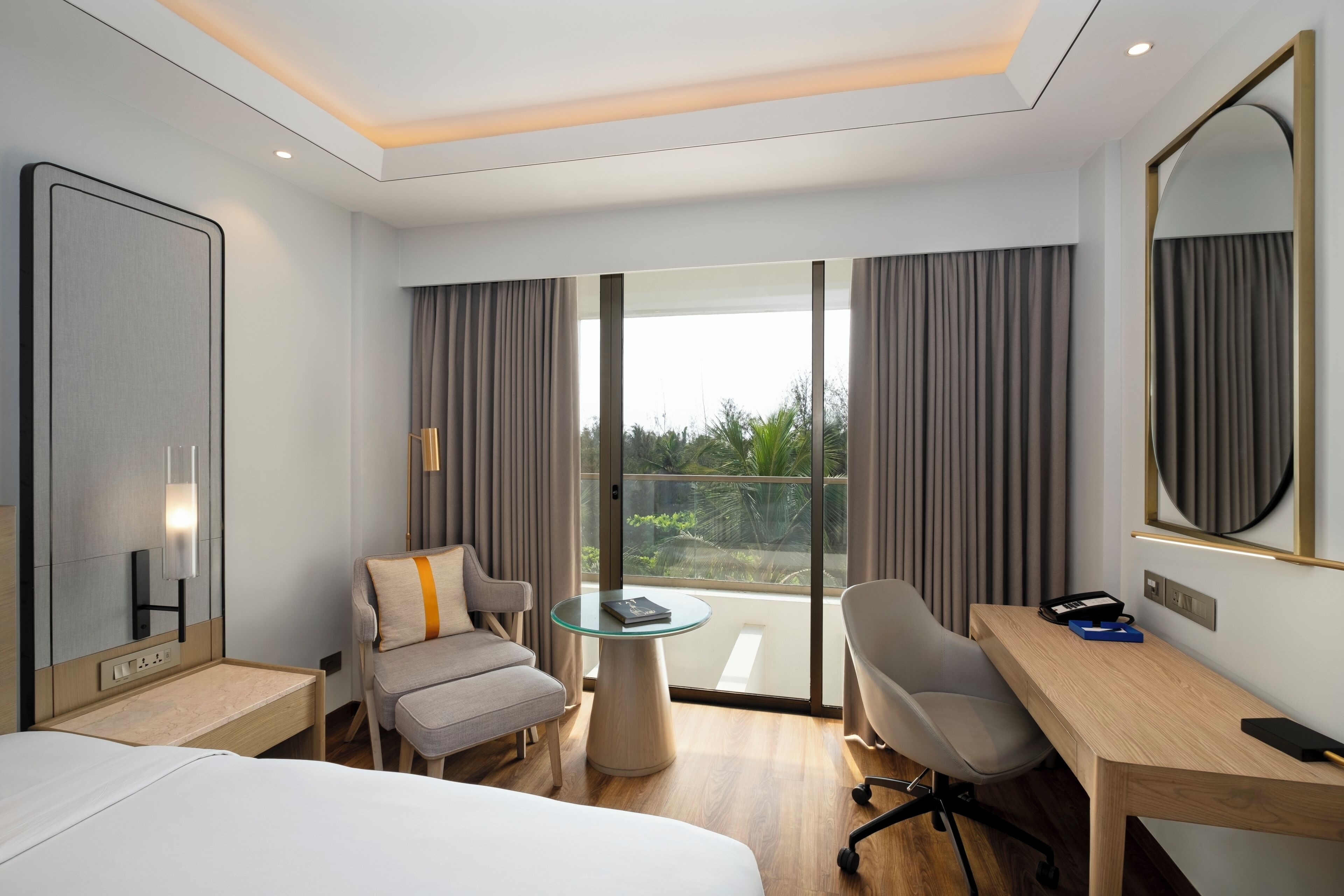 Ein modernes Hotelzimmer mit Bett, Schreibtisch, Stuhl, kleinem Tisch und einem großen Fenster mit Vorhängen.