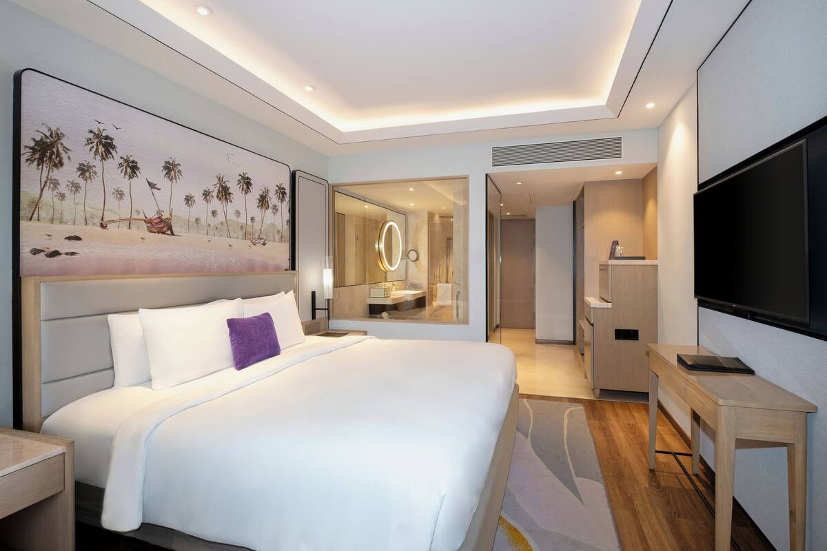 A modern hotel room with a large bed, a TV, a desk, and a bathroom visible through a glass door.