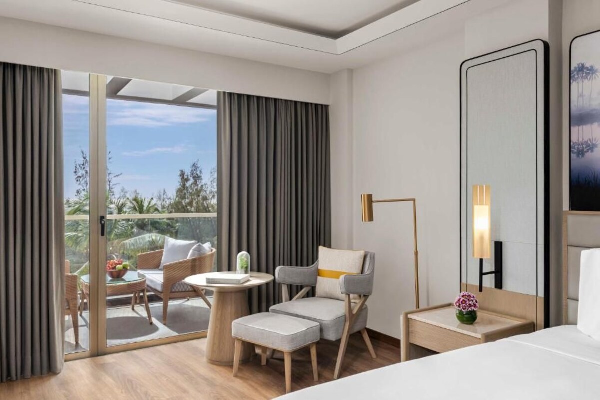 A hotel room with a balcony, a bed, a chair, a small table, and a nightstand.