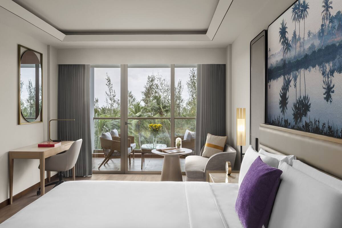 A hotel room with a large bed, a desk, a chair, a small table with a vase of flowers, and a view of palm trees through large windows.