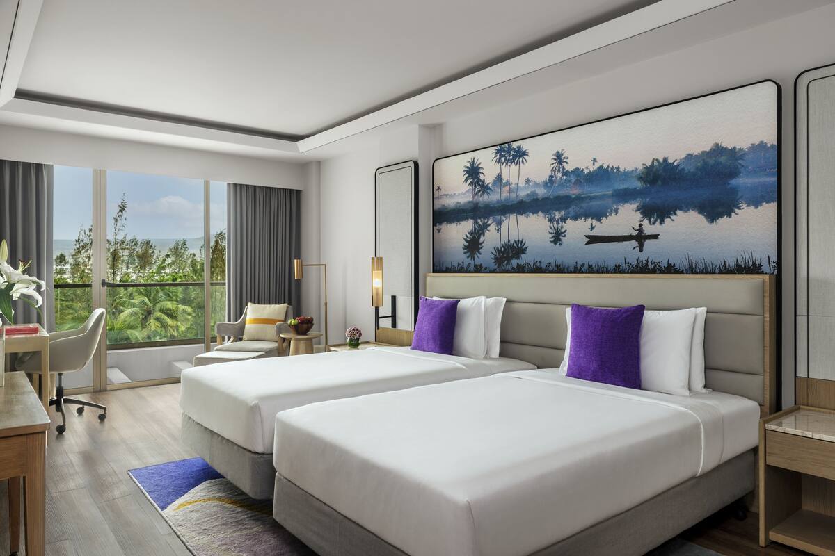 A hotel room with two beds, a desk, a chair, a large window with a view of greenery, and a painting on the wall.