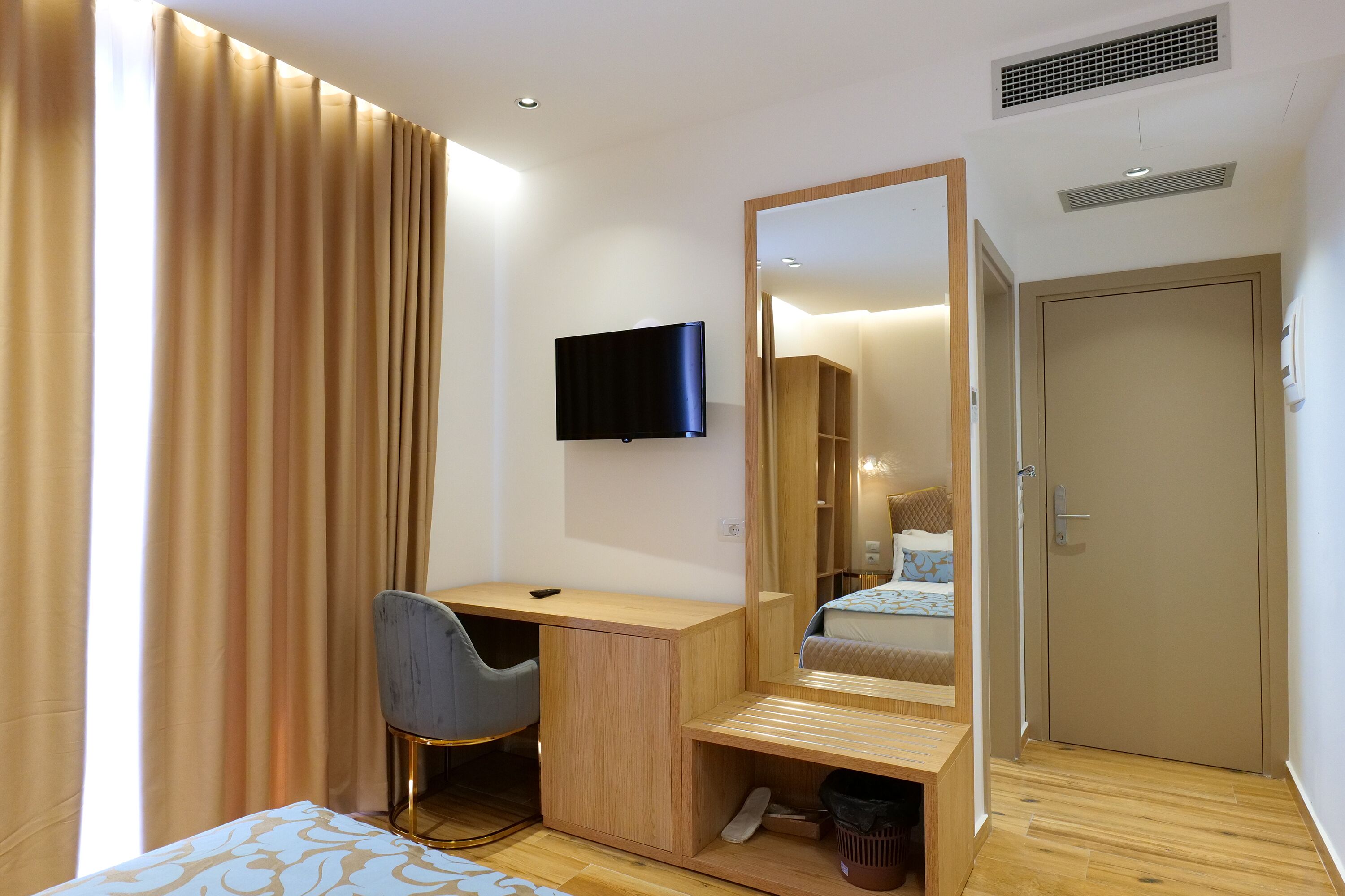 A compact hotel room with a wooden desk, a chair, a flat-screen TV, a large mirror, and a wardrobe.