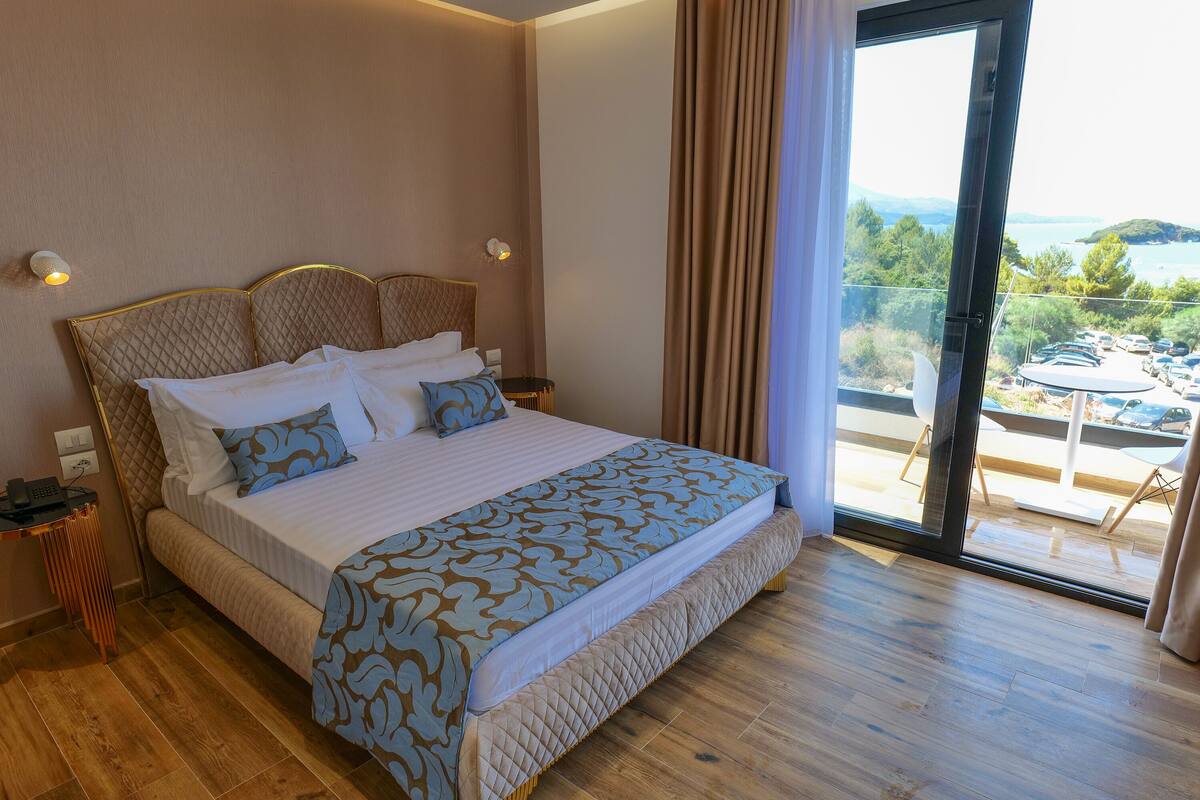Habitación de hotel con una cama grande, una mesita de noche, vista al mar y un balcón con mobiliario exterior.