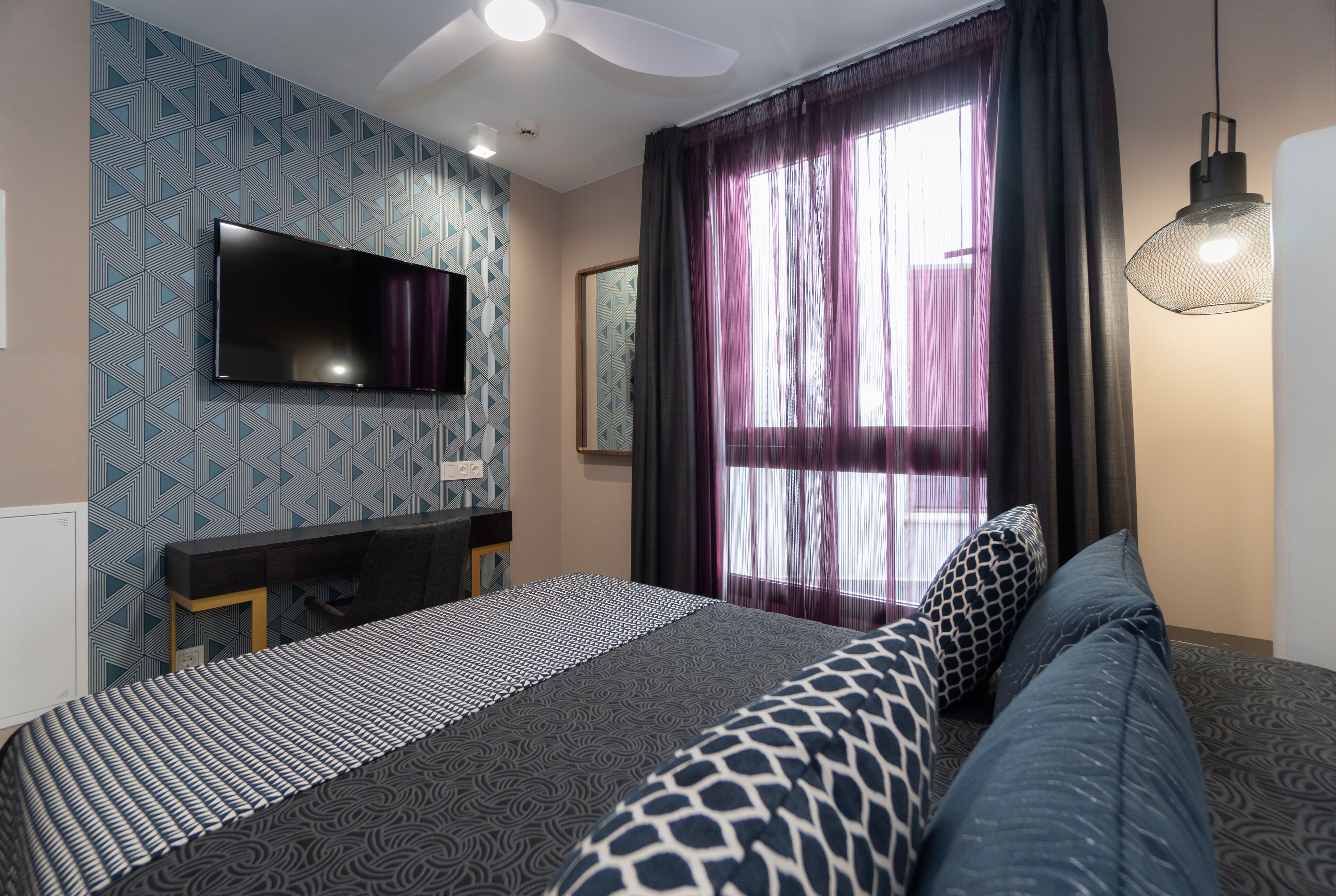 Une chambre d’hôtel moderne dotée d’un grand lit, d’un téléviseur à écran plat fixé au mur, d’un bureau avec une chaise et d’une grande fenêtre avec des rideaux.