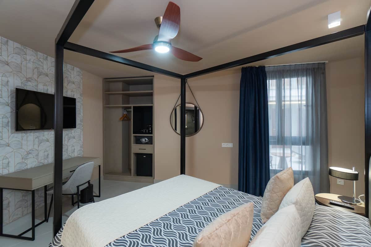 A hotel room with a bed, desk, chair, ceiling fan, and a view of the outside through a window.