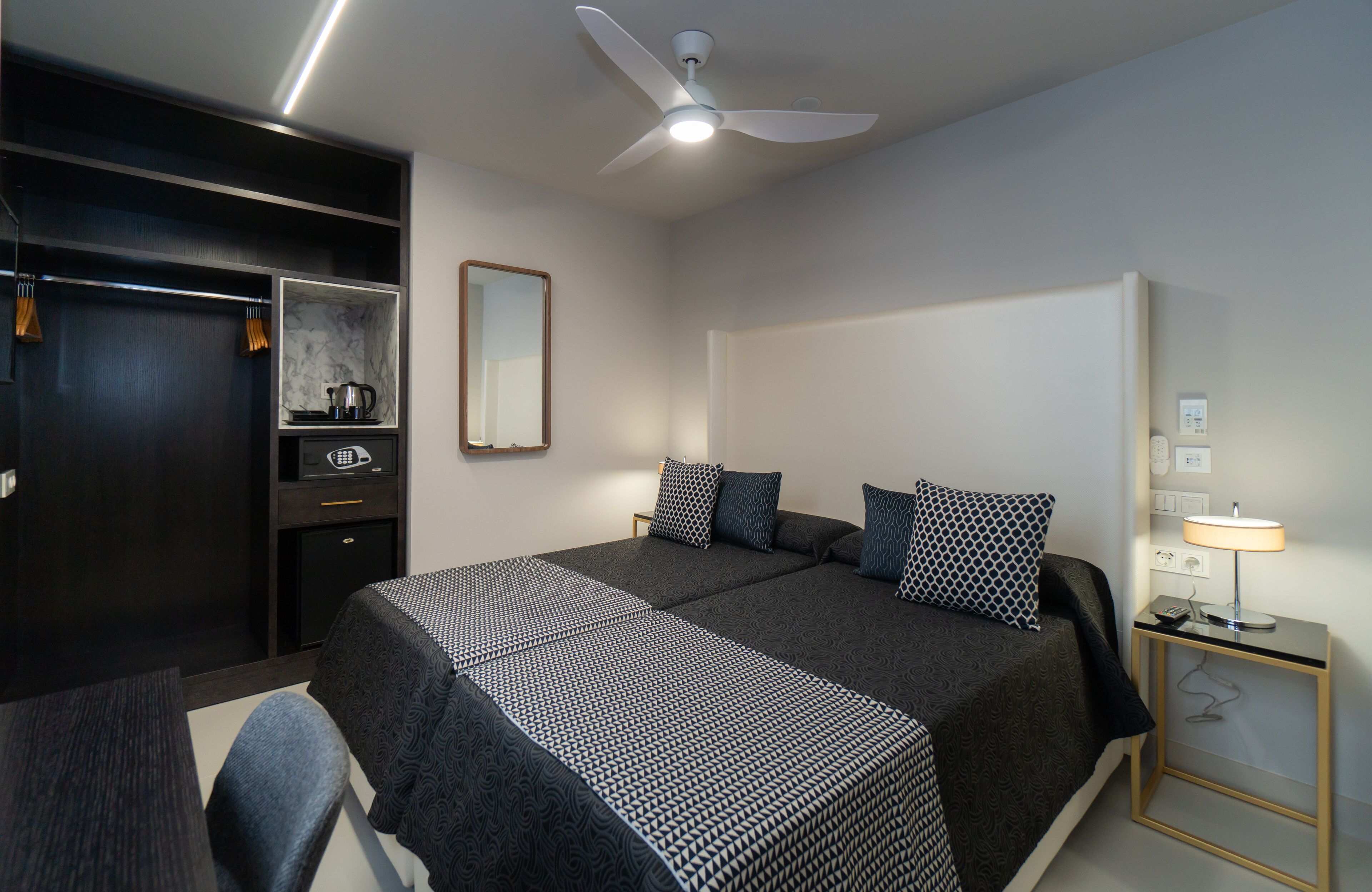 A hotel room with a large bed, a desk, a chair, a wardrobe, a mirror, a ceiling fan, and a bedside table with a lamp.