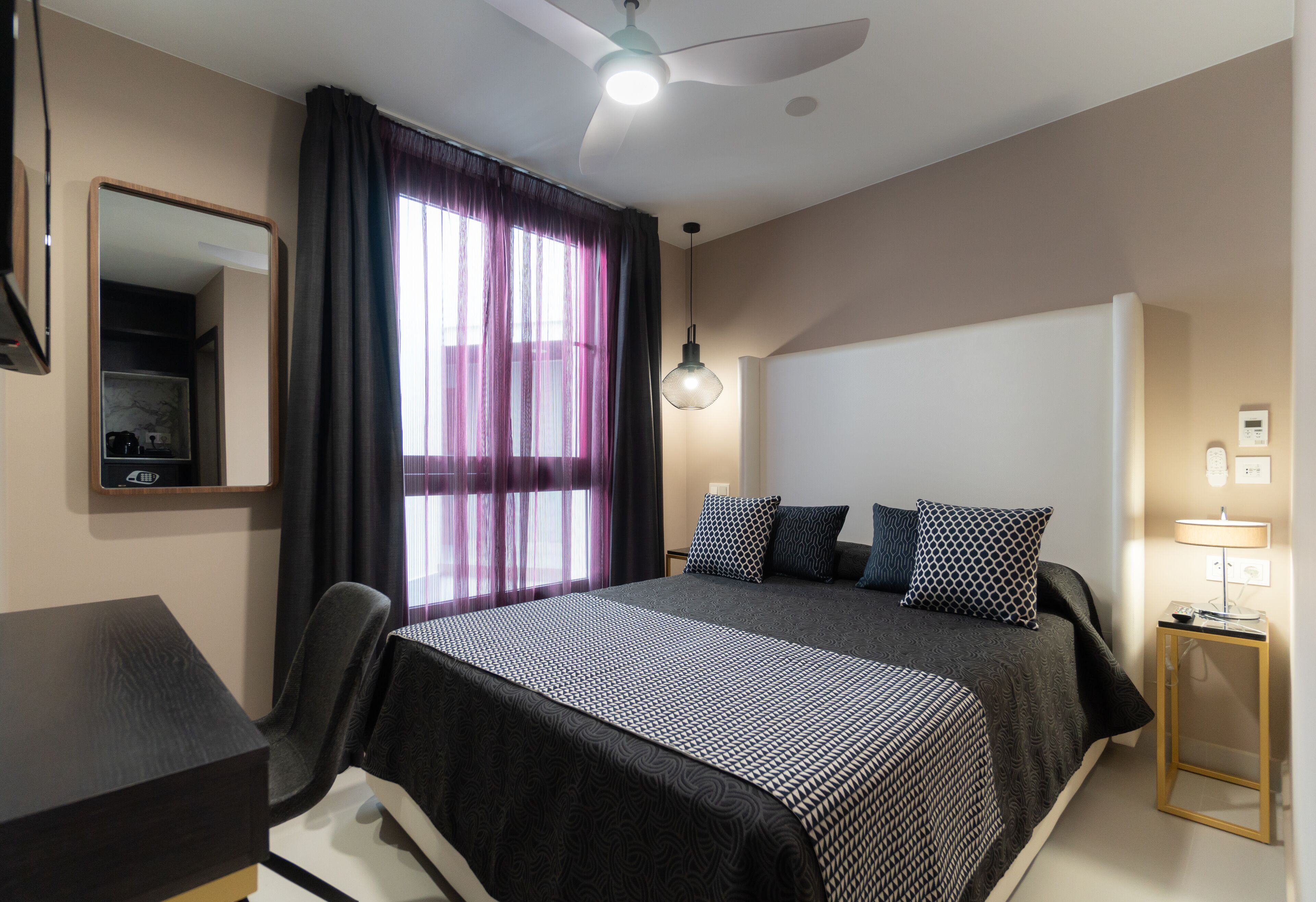 A modern hotel room with a large bed, a desk, a chair, a ceiling fan, and a window with curtains.