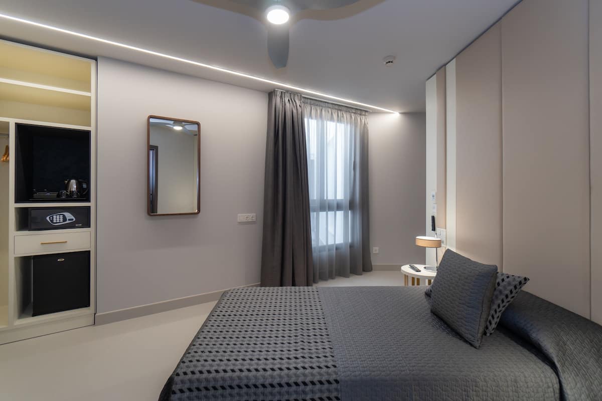 A modern hotel room with a large bed, a desk with a computer, a mirror, and a window with curtains.