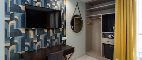 A hotel room with a bed, a desk with a chair, a TV mounted on the wall, a wardrobe, and a small closet with a mirror and hanging clothes.