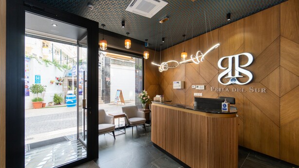 A hotel reception area with a wooden reception desk, a glass door entrance, and a view of the street outside. - Hotel Perla Del Sur (Torremolinos)