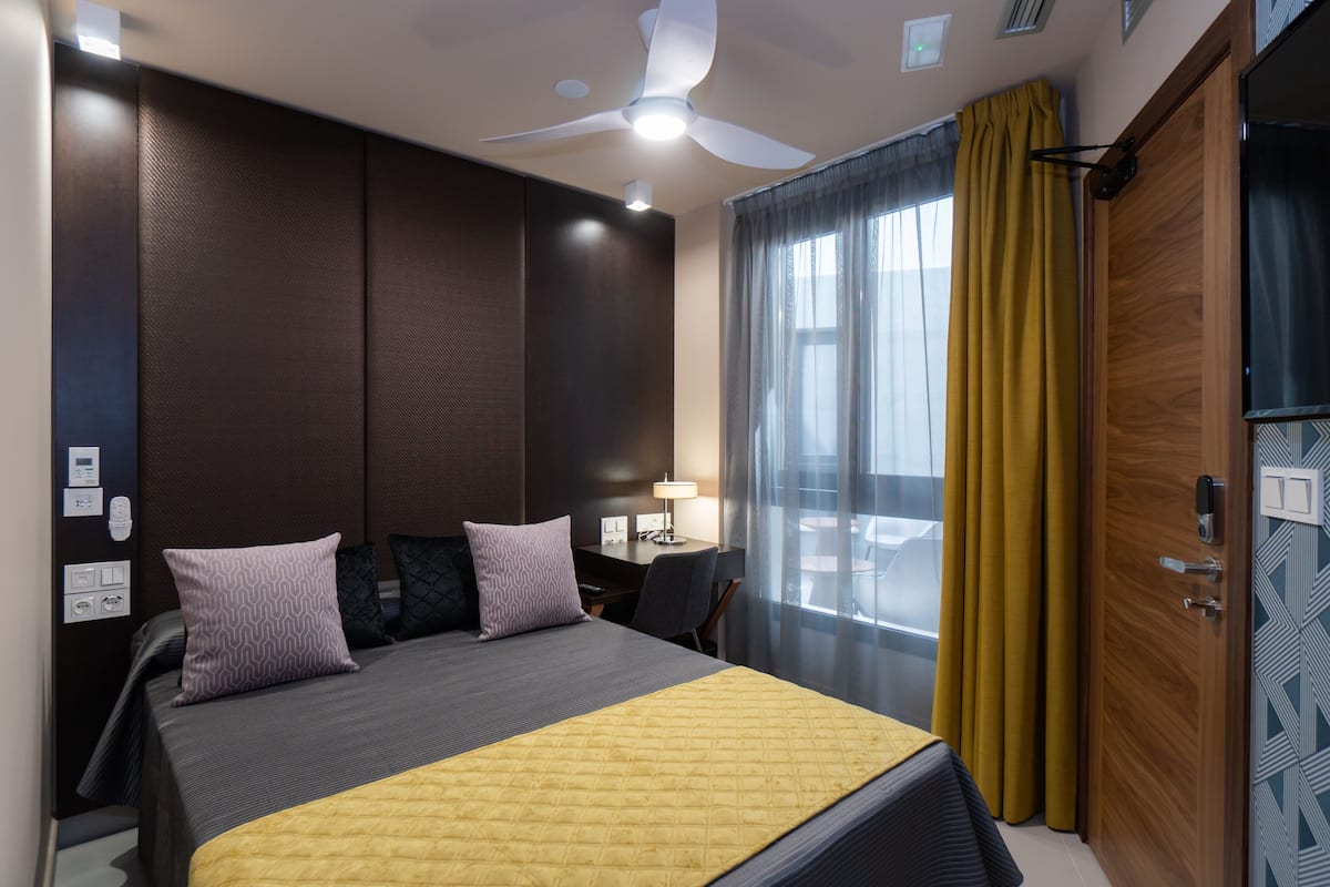 A hotel room with a bed, a desk, a chair, a desk lamp, a ceiling fan, and a window with curtains.
