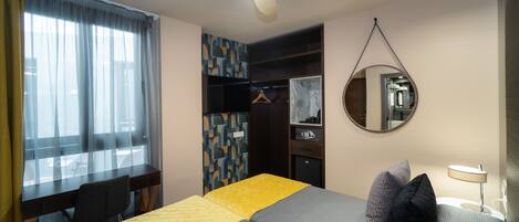 Habitación de hotel con cama, escritorio, silla, armario, espejo y ventilador de techo.
