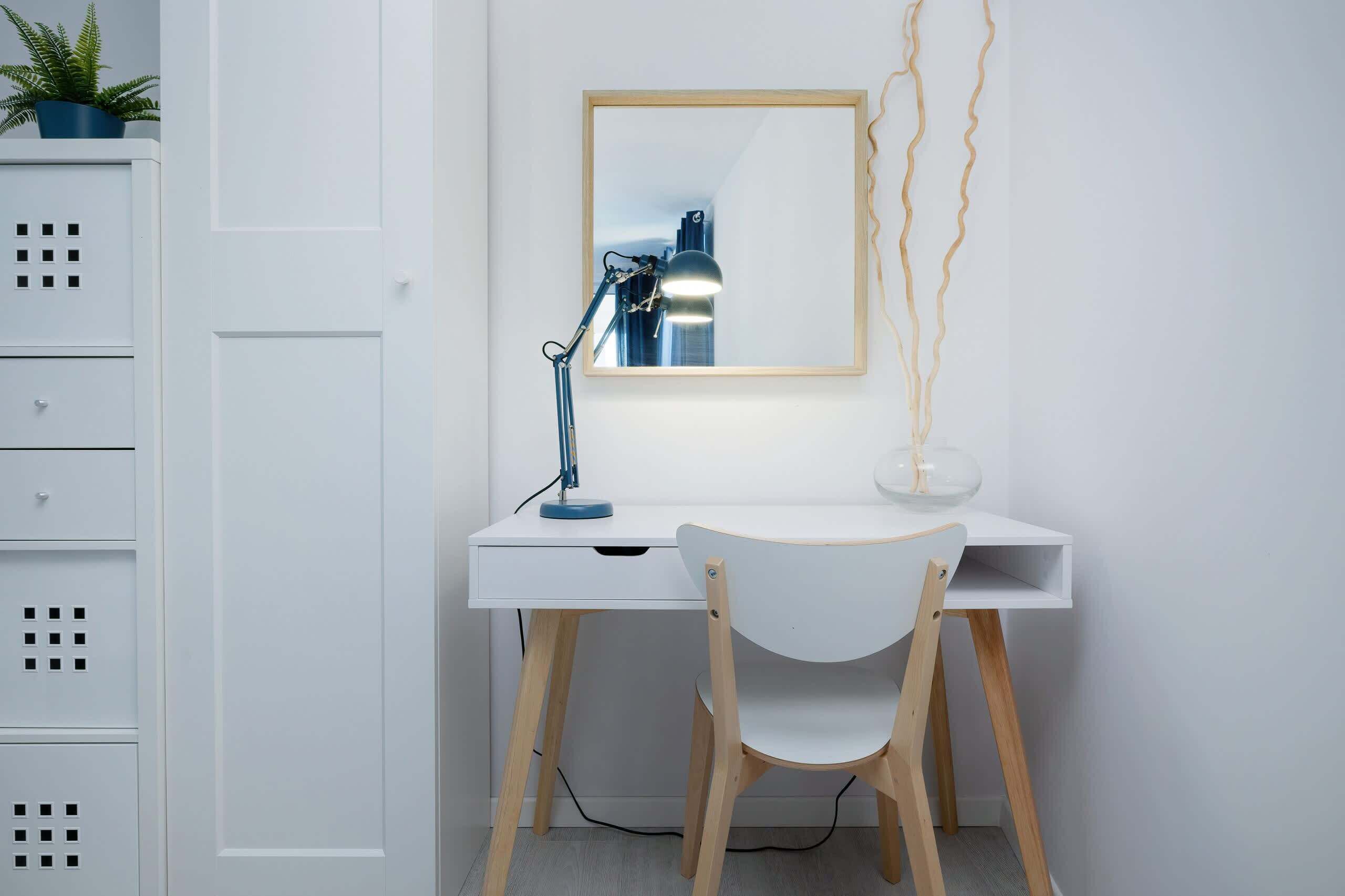 A white desk with a blue desk lamp, a white chair, and a mirror on the wall.