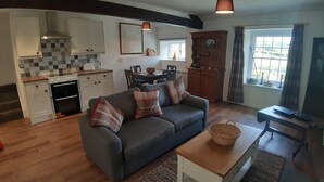 Et moderne køkken med hvide skabe, et spiseområde med et bord og stole, en grå sofa med ternede hynder og et vindue med gardiner.