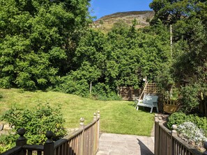 A wooden fence leading to a grassy area with a bench and a table, surrounded by dense green trees and a hill in the background.