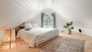 A bedroom with a sloped ceiling, a bed with white bedding, a patterned rug, a potted plant, and a floor lamp. - Beautifully renovated red cottage by the lake (Salo)