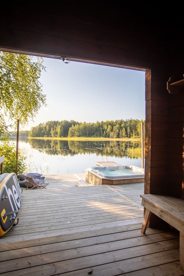 A wooden deck with a hot tub and a boat docked on a calm lake, surrounded by trees.