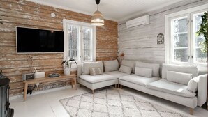 A modern living room with a white sectional sofa, a flat-screen TV mounted on a wooden wall, and a hanging pendant light. - Beautifully renovated red cottage by the lake (Salo)