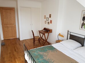 Quarto com uma cama, uma escrivaninha com cadeira, um abajur e uma porta.