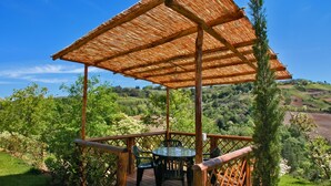 Une terrasse en bois avec une table et des chaises, entourée d’une végétation luxuriante et avec des collines en arrière-plan.