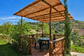 A wooden deck with a table and chairs, surrounded by lush greenery and hills in the background.