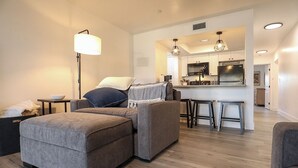 Una sala de estar moderna con un sofá seccional gris, una lámpara de pie y una cocina visible a través de un umbral abierto.
