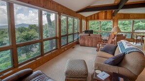 Amplia habitación con ventanas grandes que ofrecen vista a un área boscosa. Se observa un sofá, una silla y una pequeña mesa con una lámpara.