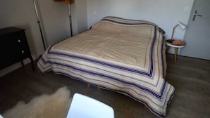 A bedroom with a sloped ceiling, a bed with a striped bedspread, a hanging light fixture, and a small table with a chair. - Comfortable house in Portnavalo by the sea (Arzon)