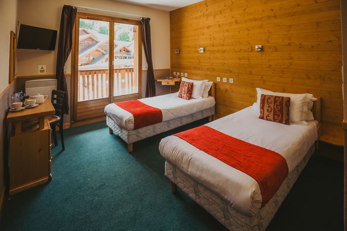 Habitación de hotel con dos camas, un escritorio con silla, televisor montado en la pared y vistas a una terraza de madera y edificios.