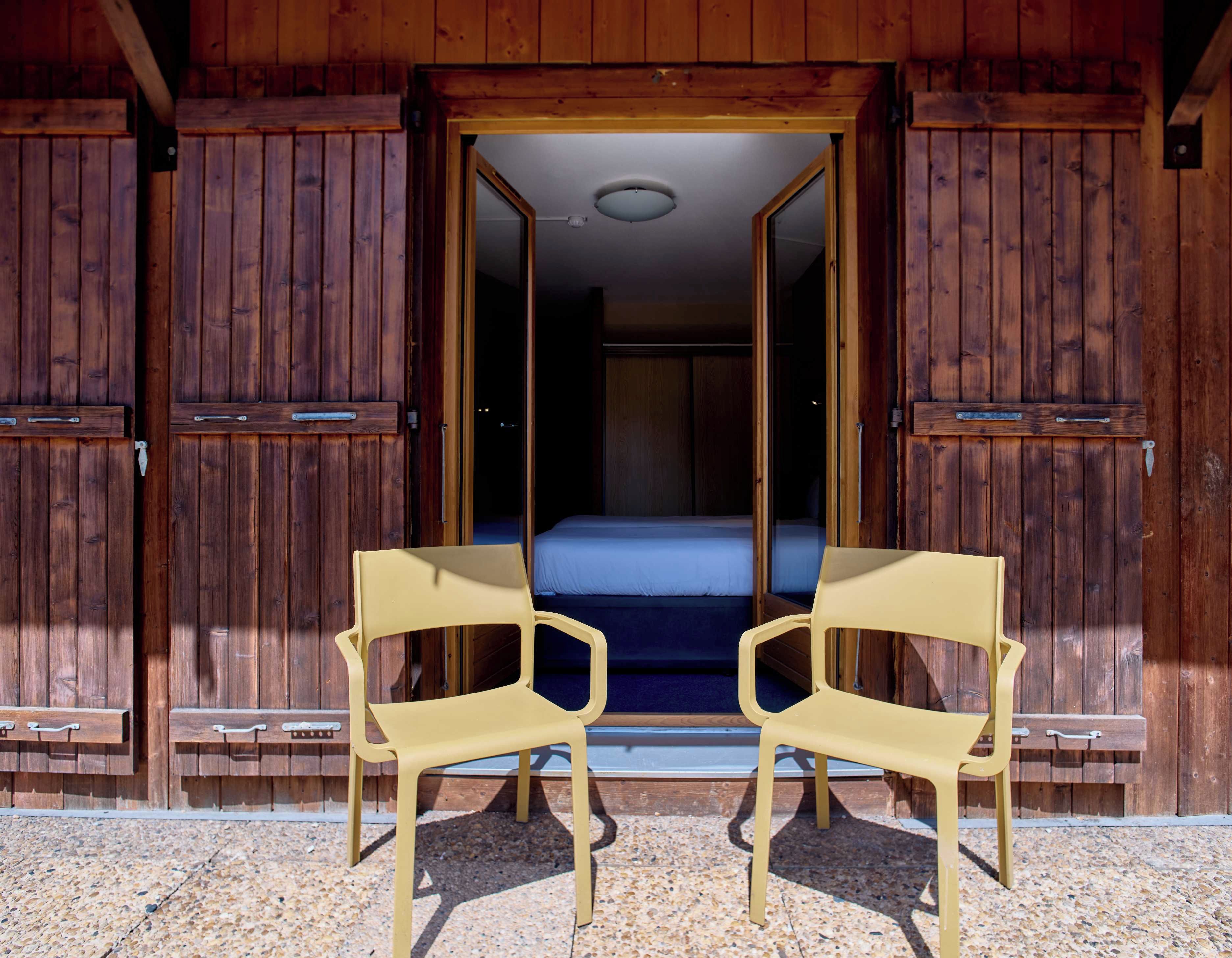 Un bâtiment en bois avec une porte ouverte donnant sur une pièce où se trouvent un lit et deux chaises.