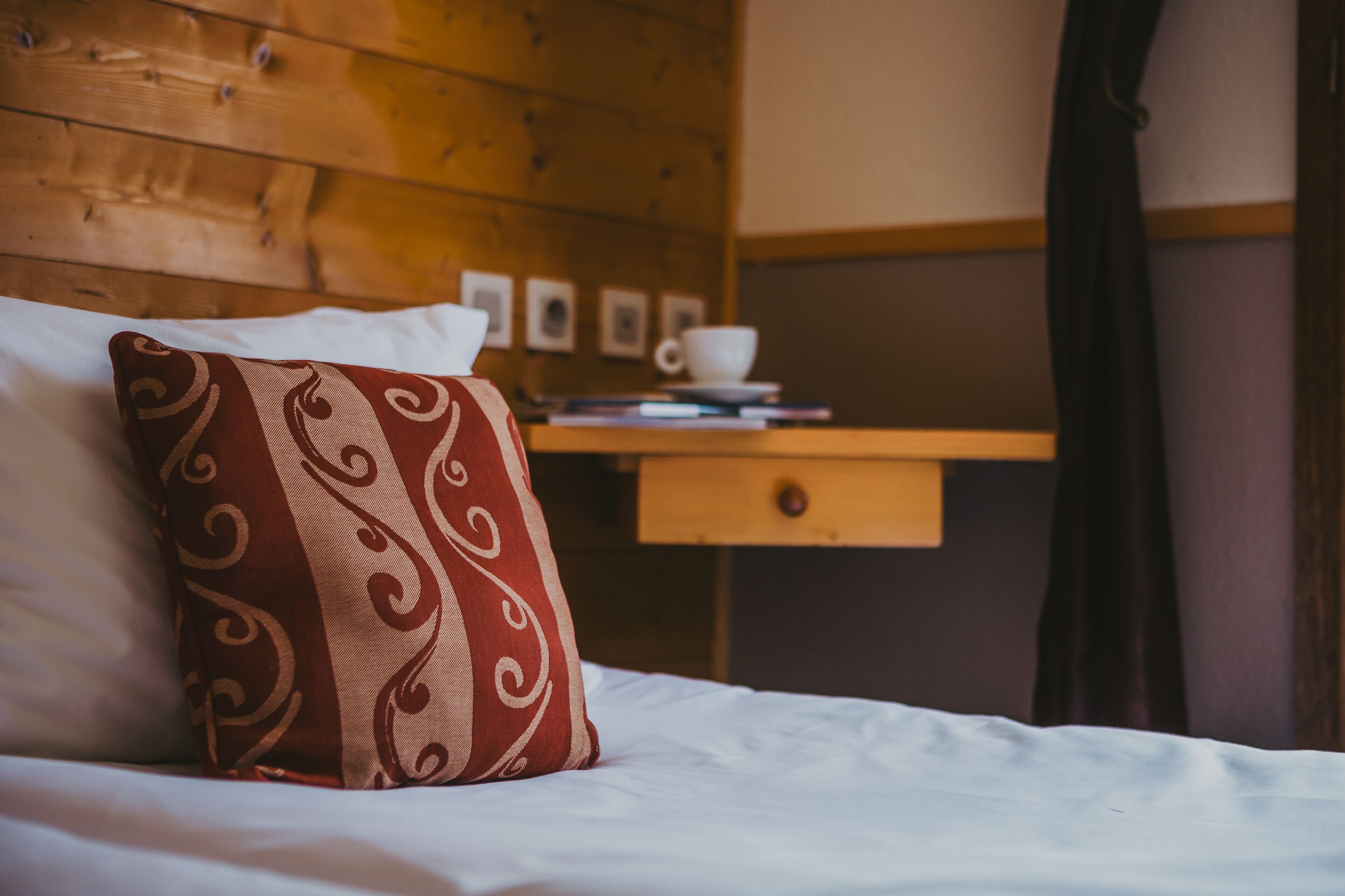 Ein ordentlich bezogenes Bett mit einem dekorativen Kissen, ein Holzkopfbrett, ein kleiner Nachttisch mit Tasse und Büchern sowie eine Wand mit gerahmten Bildern.