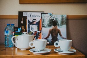 Una mesa de madera con dos tazas blancas, una tetera, una botella de agua y revistas.