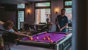 Une table de billard dans une pièce dotée de chaises et de tables, de grandes fenêtres et de luminaires fixés au mur.