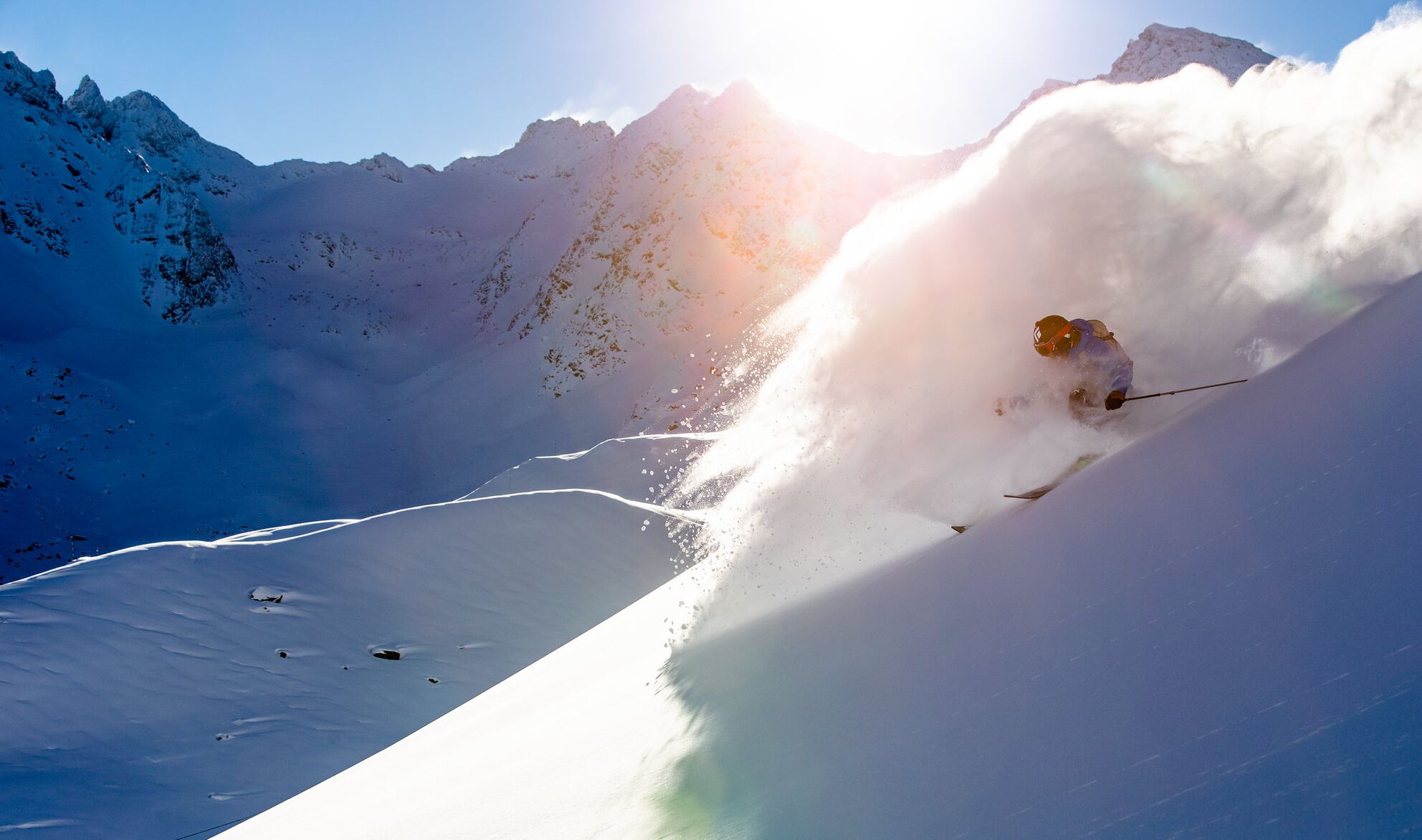 Un esquiador descendiendo una pendiente pronunciada cubierta de nieve, bajo un cielo despejado y con montañas a la distancia.