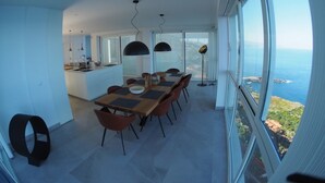 Un moderno comedor con una gran mesa y sillas, una cocina al fondo y una vista panorámica del océano a través de amplios ventanales.