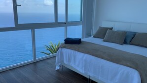 Un dormitorio con un ventanal grande con vistas al mar, una cama con cubrecama blanco y palmeras a la vista.